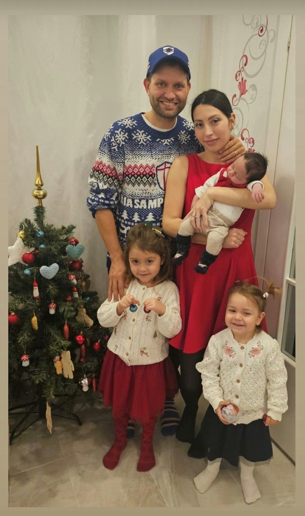 Eine Familie mit drei kleinen Kindern steht neben einem geschmückten Weihnachtsbaum und lacht in die Kamera. Die Mutter hält ein Baby im Arm, während die beiden älteren Kinder Weihnachtskugeln in den Händen halten.