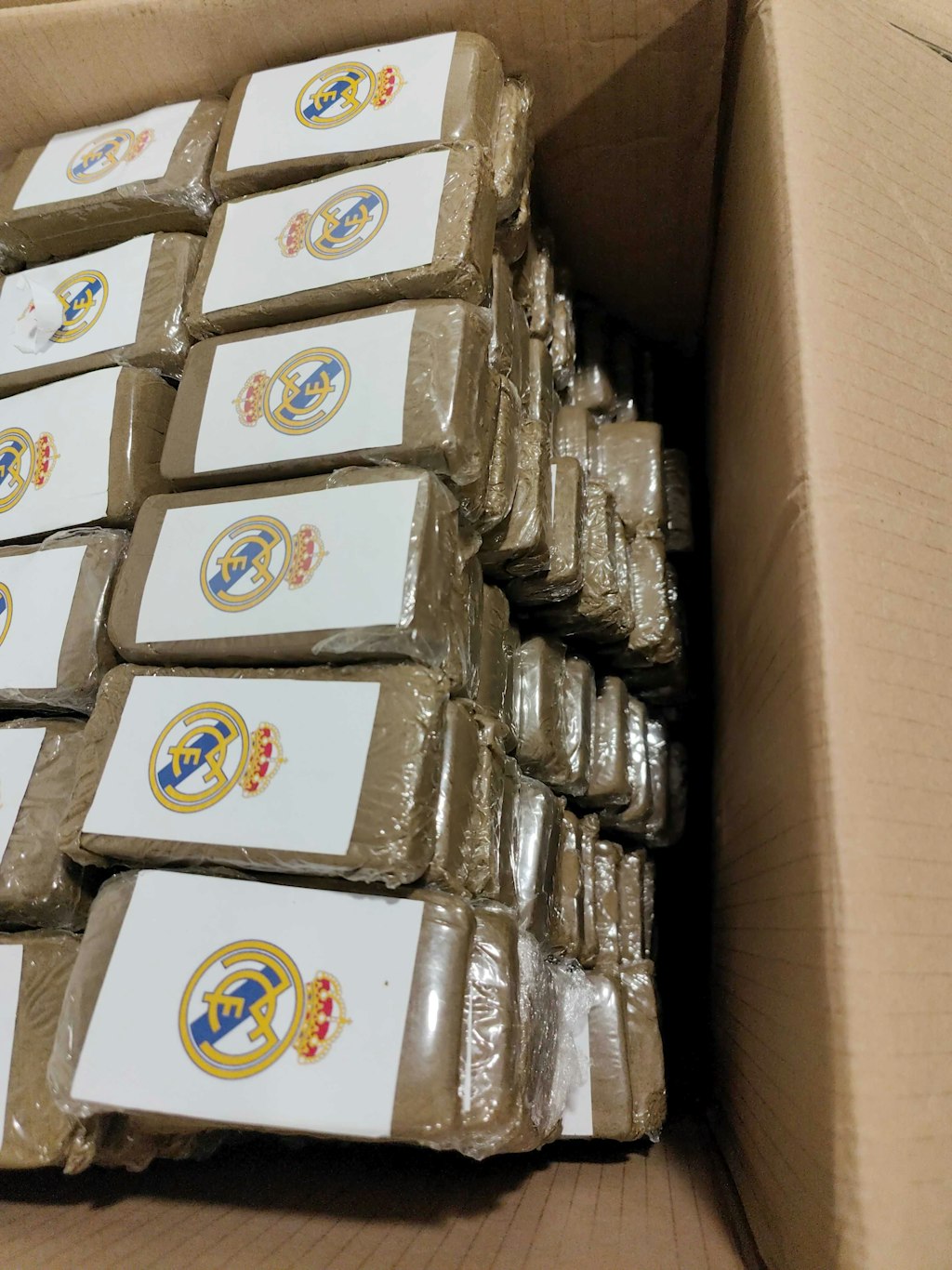 In einem Karton befinden sich zahlreiche Pakete, die mit einem Logo versehen sind. Diese Pakete enthalten CBD mit synthetischen Cannabinoiden. Die Verpackungen sind ordentlich gestapelt.