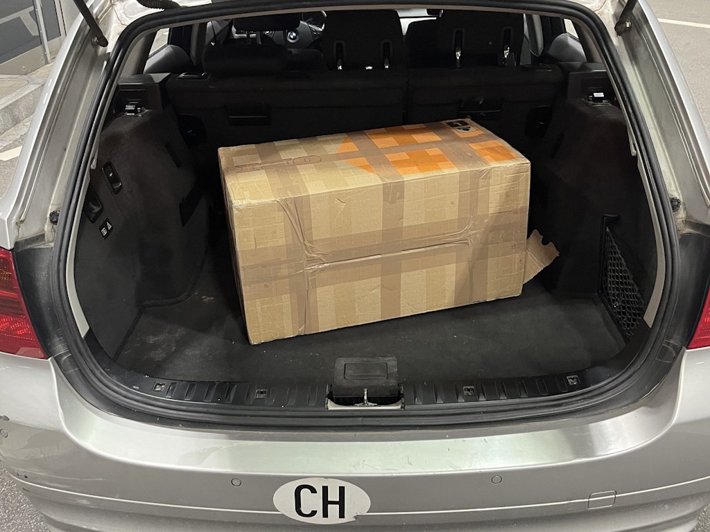 Eine Kartonkiste befindet sich im Kofferraum eines Autos. Der Kofferraum ist geöffnet und das Auto trägt ein CH-Kennzeichen.