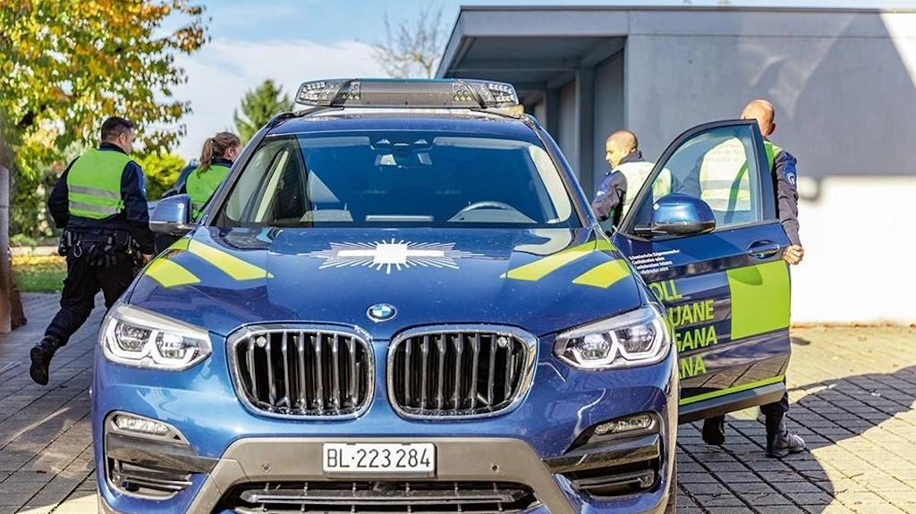 Ein blaues Zollfahrzeug steht mit geöffneter Tür auf einem Parkplatz. Mehrere Mitarbeitende des BAZG in Uniformen sind um das Fahrzeug herum zu sehen.