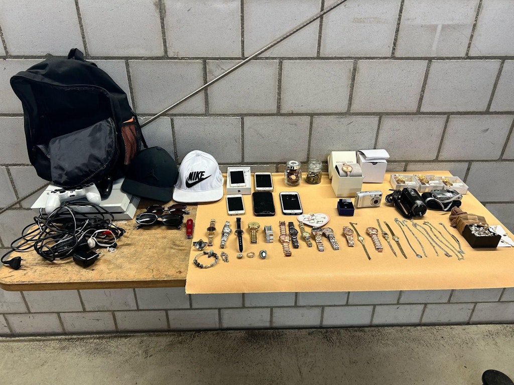 Eine Sammlung von Schmuck, Elektronikgeräten und Accessoires auf einem Tisch in einem Raum mit grauen Wänden.