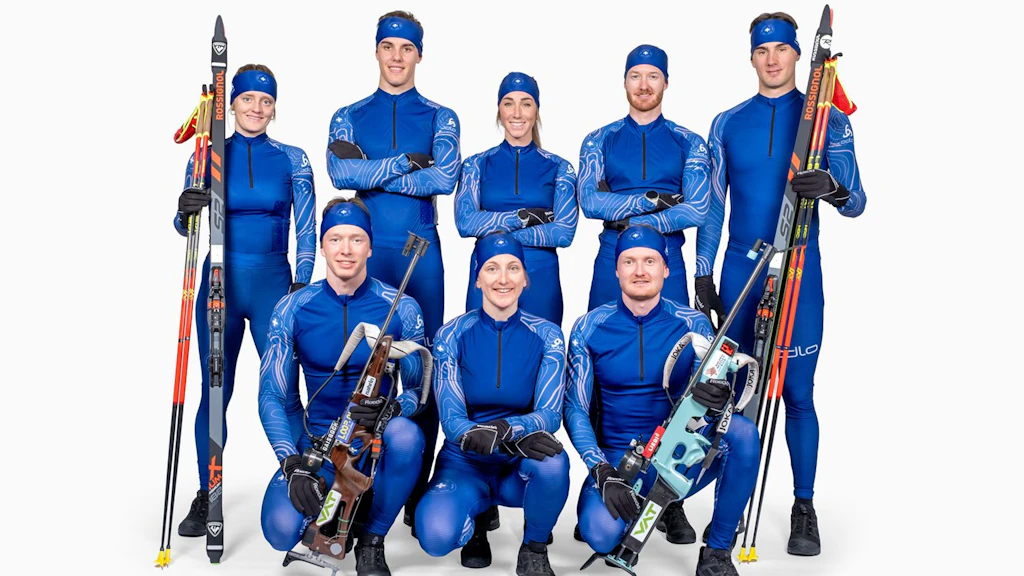 Eine Gruppe von neun Personen in blauen Sportanzügen posiert mit Biathlon-Ausrüstung. Sie tragen Skier und Gewehre und lächeln in die Kamera.