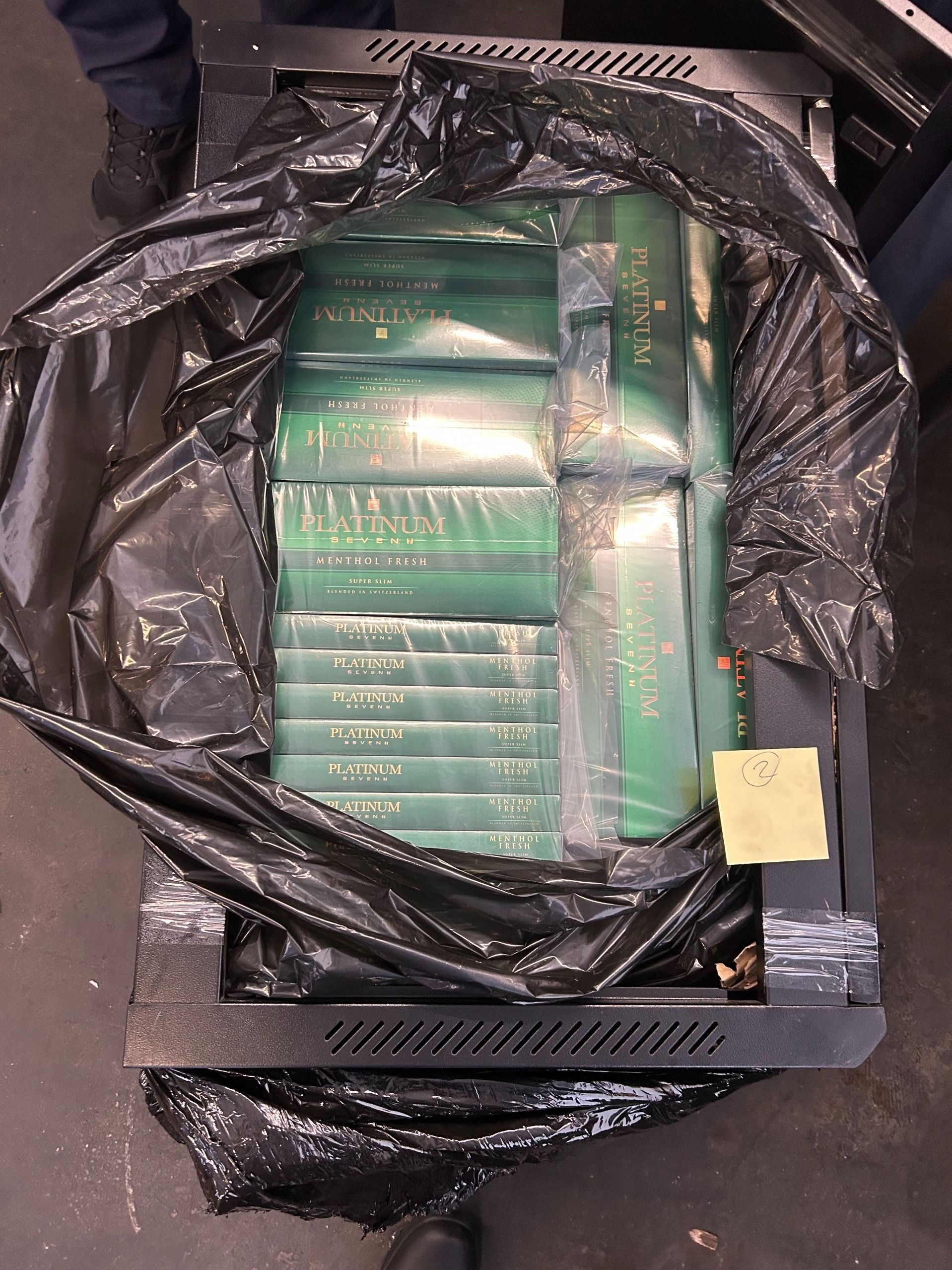 In einem geöffneten Computerrack sind zahlreiche grüne Zigarettenstangen der Marke Platinum versteckt. Die Zigaretten sind in einer schwarzen Plastiksacke eingewickelt und sorgfältig im Rack platziert.