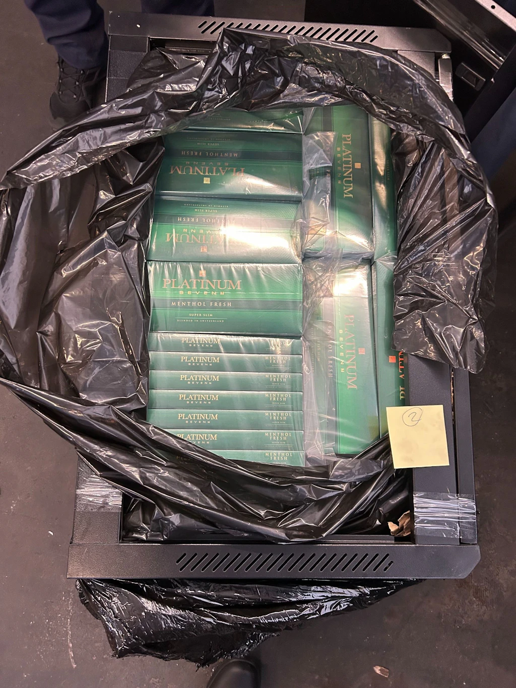 In einem geöffneten Computerrack sind zahlreiche grüne Zigarettenstangen der Marke Platinum versteckt. Die Zigaretten sind in einer schwarzen Plastiksacke eingewickelt und sorgfältig im Rack platziert.