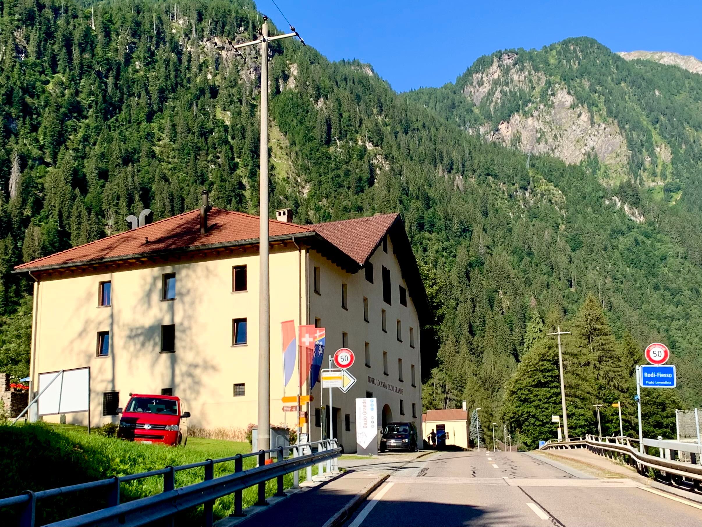Ein Hotelgebäude in einer bergigen Landschaft mit einem roten Auto davor. Schilder und Fahnen sind ebenfalls sichtbar.