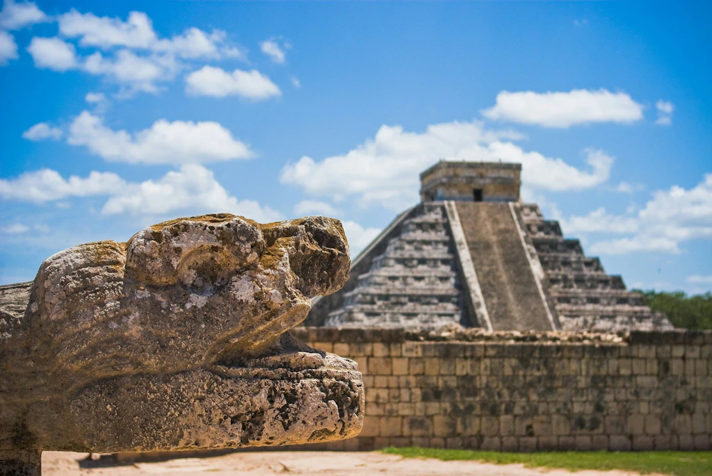 Im Vordergrund ist eine steinerne Skulptur eines Tieres zu sehen, im Hintergrund steht eine grosse Stufenpyramide unter blauem Himmel mit Wolken. Die Szene wirkt historisch und monumental.