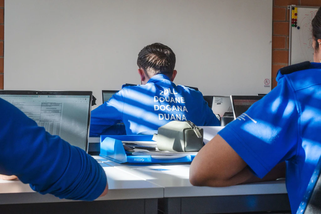 Mehrere Zollbeamte in blauer Uniform sitzen an Computern in einem Schulungsraum. Auf dem Rücken eines Beamten stehen die Begriffe Zoll, Douane, Dogana und Duana. Die Szene zeigt eine Lernsituation im Innenraum.  