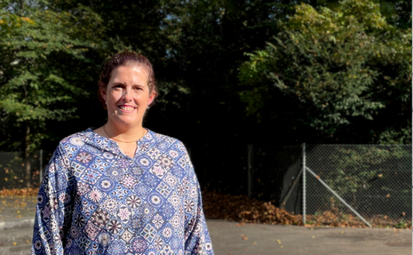 Caroline von Riedmatten steht lächelnd im Freien vor einem Zaun und herbstlicher Vegetation. Sie trägt eine gemusterte Bluse.