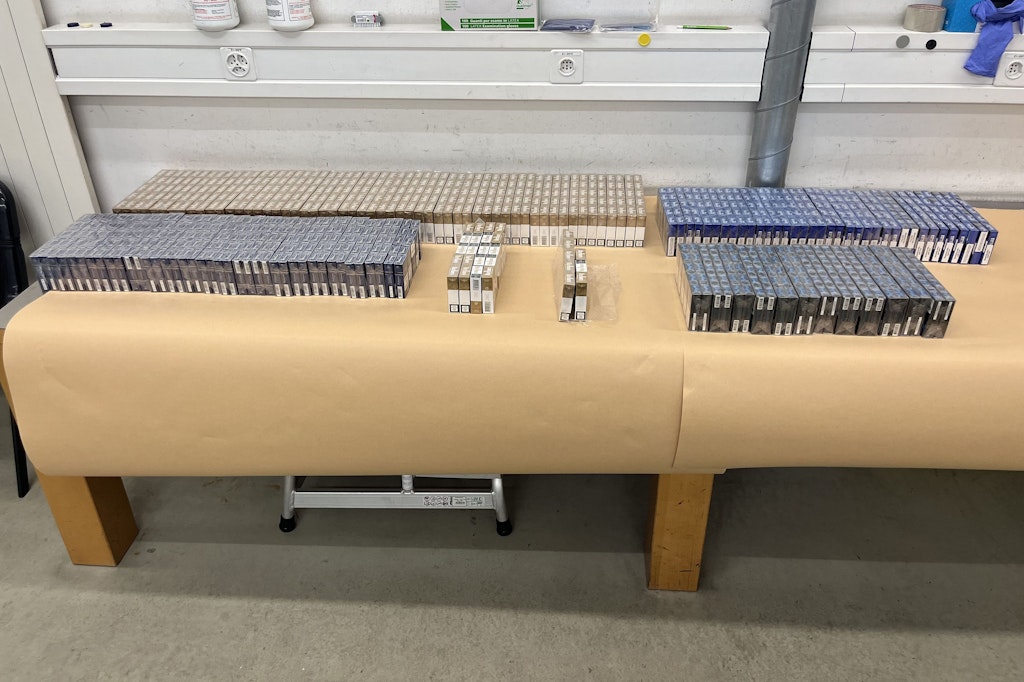 Eine grosse Menge an Zigarettenpackungen ist auf einem Tisch ausgebreitet. Die Packungen sind in verschiedenen Farben und Grössen sortiert. Der Tisch steht in einem Raum mit weissen Wänden und Regalen.