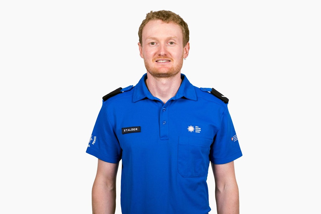 Ein Mann in einer blauen Zolluniform mit Namensschild "Stalder". Er trägt eine Uniform mit der Aufschrift "Zoll Douane Dogana Duana".