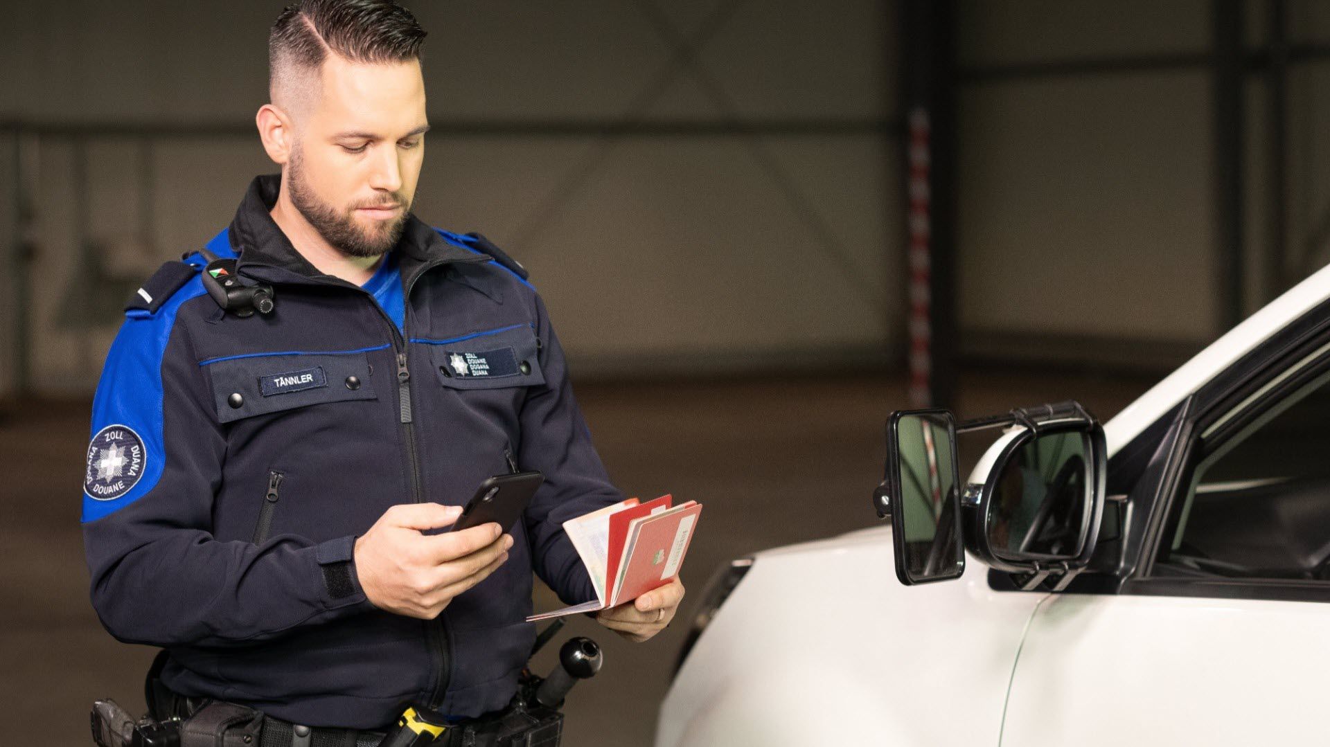 Ein Zollmitarbeiter überprüft Pässe und ein Mobiltelefon neben einem Fahrzeug. 