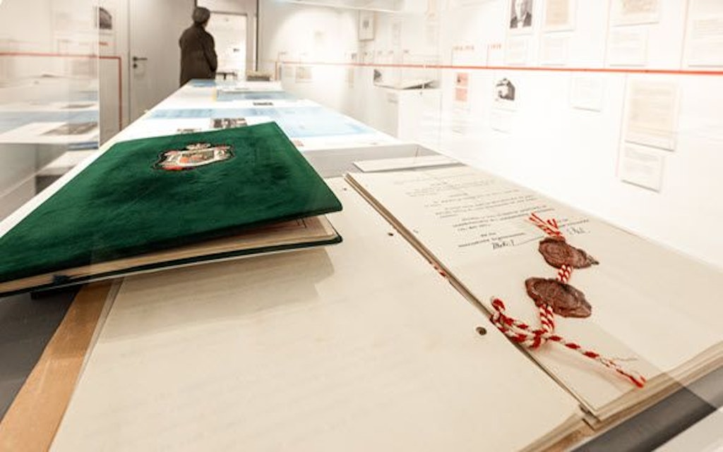 Eine Ausstellung im Schweizer Zollmuseum zeigt historische Dokumente zum 100-jährigen Zollvertrag zwischen der Schweiz und Liechtenstein. Im Vordergrund sind ein Buch mit einem Wappen und ein versiegeltes Dokument zu sehen.