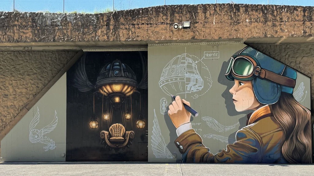 Ein Wandgemälde zeigt eine Pilotin mit Fliegerbrille, die ein futuristisches Luftschiff entwirft. Daneben ist ein beleuchteter Sessel in einem dunklen Raum zu sehen.  