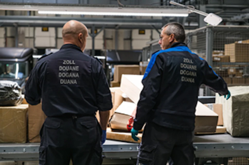 Zwei Mitarbeitende des BAZG stehen an einem Förderband mit Paketen und kontrollieren Sendungen. Sie tragen Uniformen mit der Aufschrift "Zoll Douane Dogana Duana" auf dem Rücken. Die Szene spielt in einer Lagerhalle oder einem Logistikzentrum.