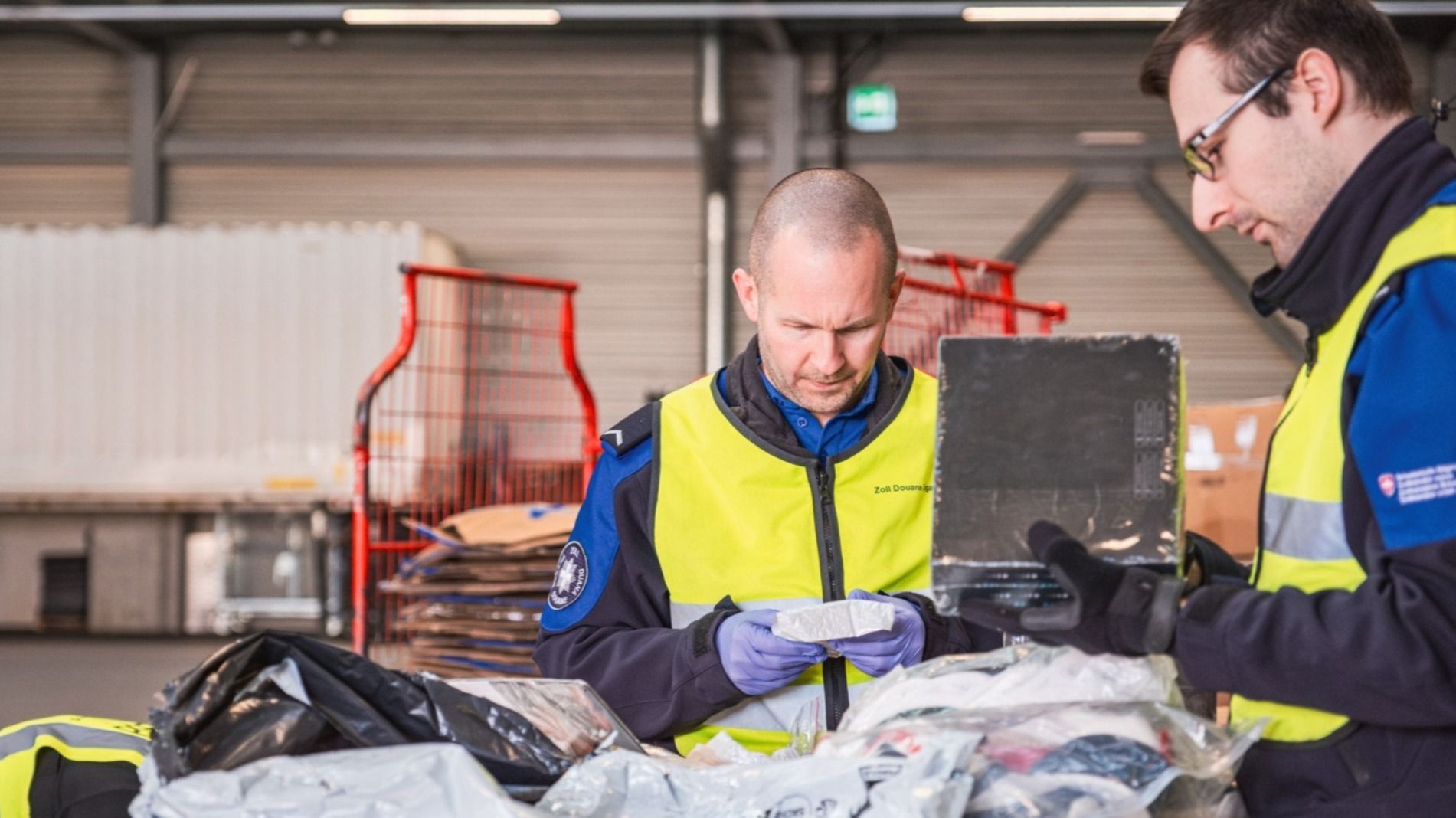Zwei Zollmitarbeiter überprüfen Pakete in einem Lager. Sie tragen Uniformen mit der Aufschrift "Zoll" und stehen vor einem Förderband voller Pakete.