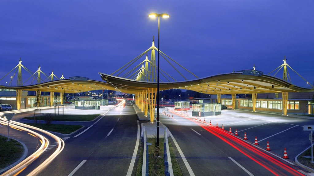 Zeitrafferaufnahme eines Autobahn Grenzübergangs bei Nacht. Die Autobahn zeigt zwei Spuren in jede Richtung, mit sichtbaren weißen und roten Lichtstreifen der Autos, die Bewegung suggerieren. Der beleuchtete Grenzübergang verfügt über ein Dach, unter dem sich pro Fahrtrichtung zwei Kabinen befinden. Neben der Autobahn sind durch reflektierende Verkehrsleitkegel abgetrennte Lastwagenparkplätze erkennbar.