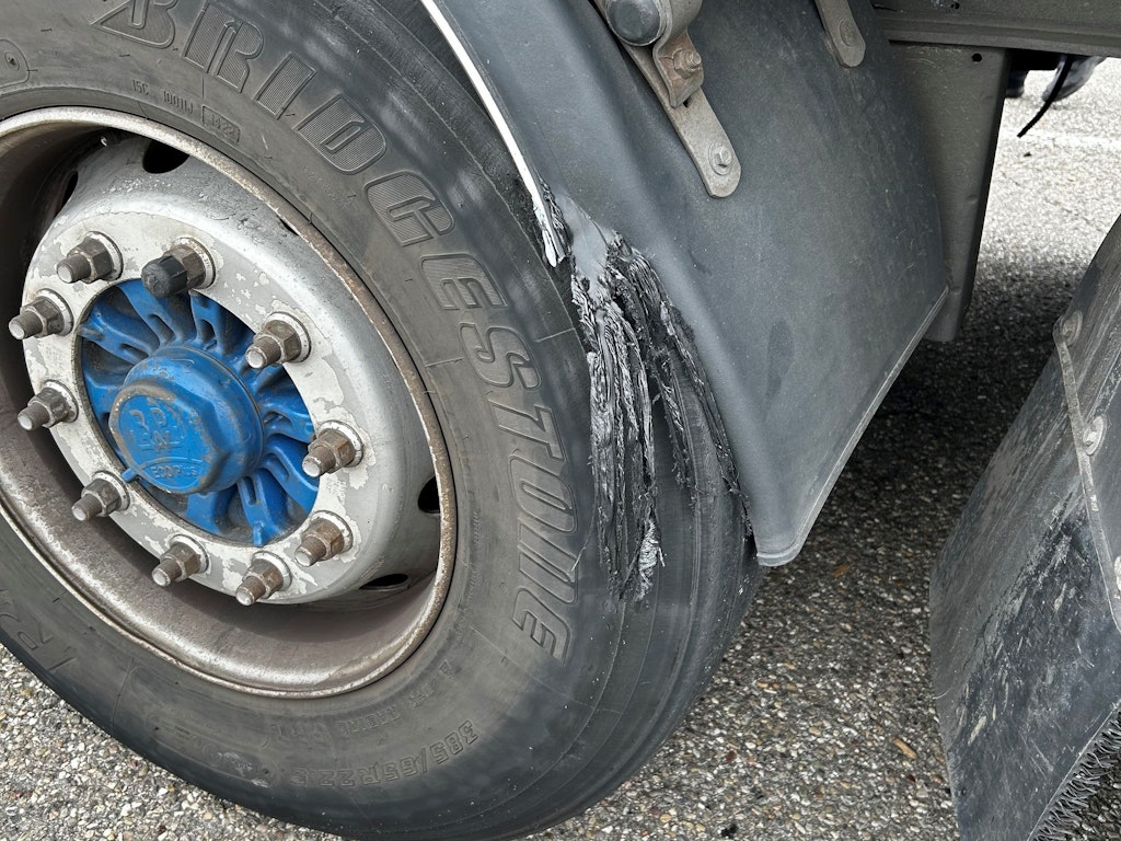 Ein LKW-Reifen mit einem beschädigten Kotflügel. Der Reifen zeigt Abnutzungsspuren, und der Kotflügel hat sichtbare Kratzer und Dellen.