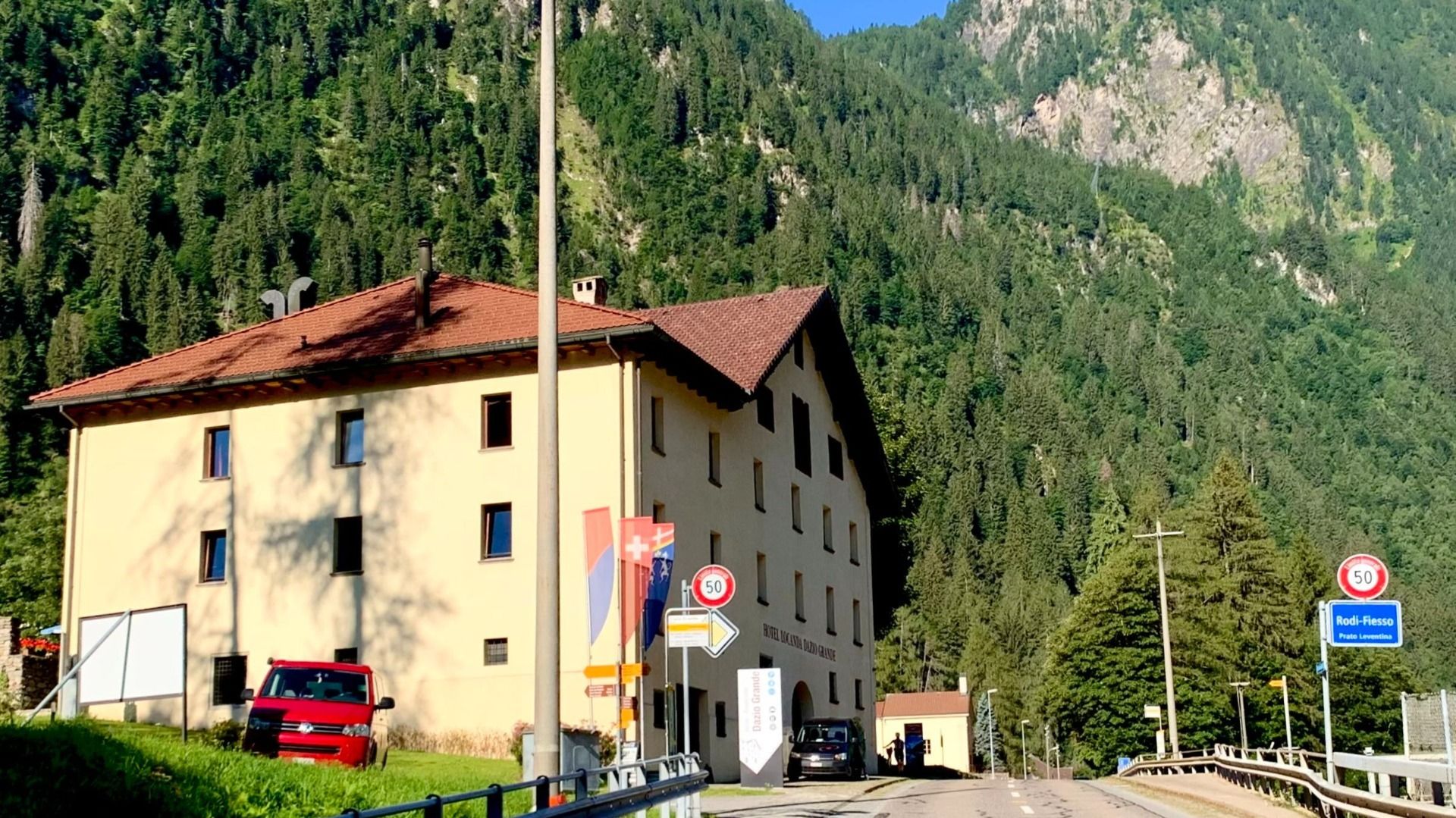 Ein Hotelgebäude in einer bergigen Landschaft mit einem roten Auto davor. Schilder und Fahnen sind ebenfalls sichtbar.  