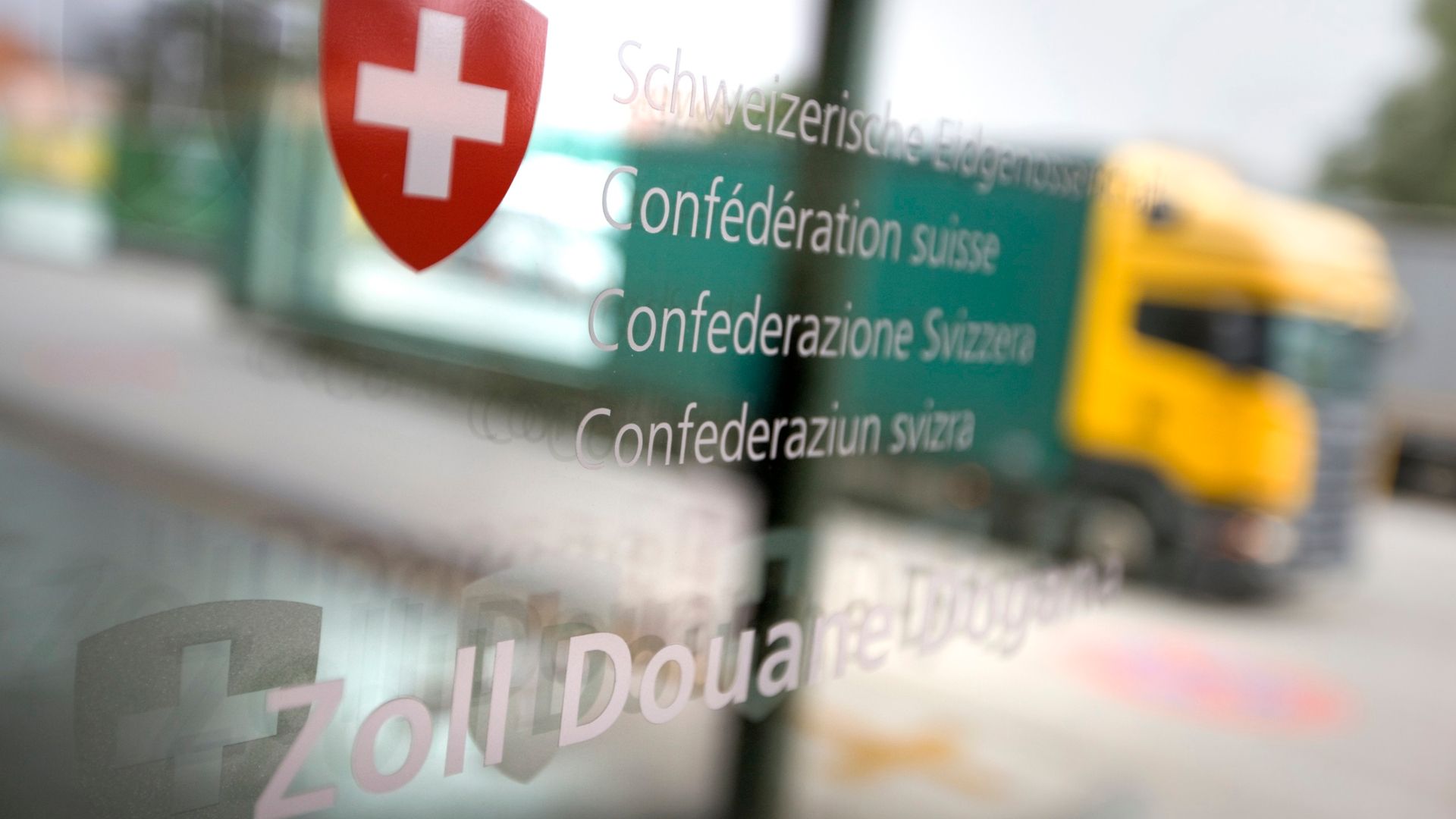 Glastür mit dem Schweizer Kreuz sowie dem Schriftzug Schweizerische Eidgenossenschaft, Confédération suisse, Confederazione Svizzera, Confederaziun svizra, Zoll, Douane, Dogana. In der Tür spiegelt sich die Strasse und ein Lastwagen mit einer gelben Führerkabine und einem grünen Auflieger.