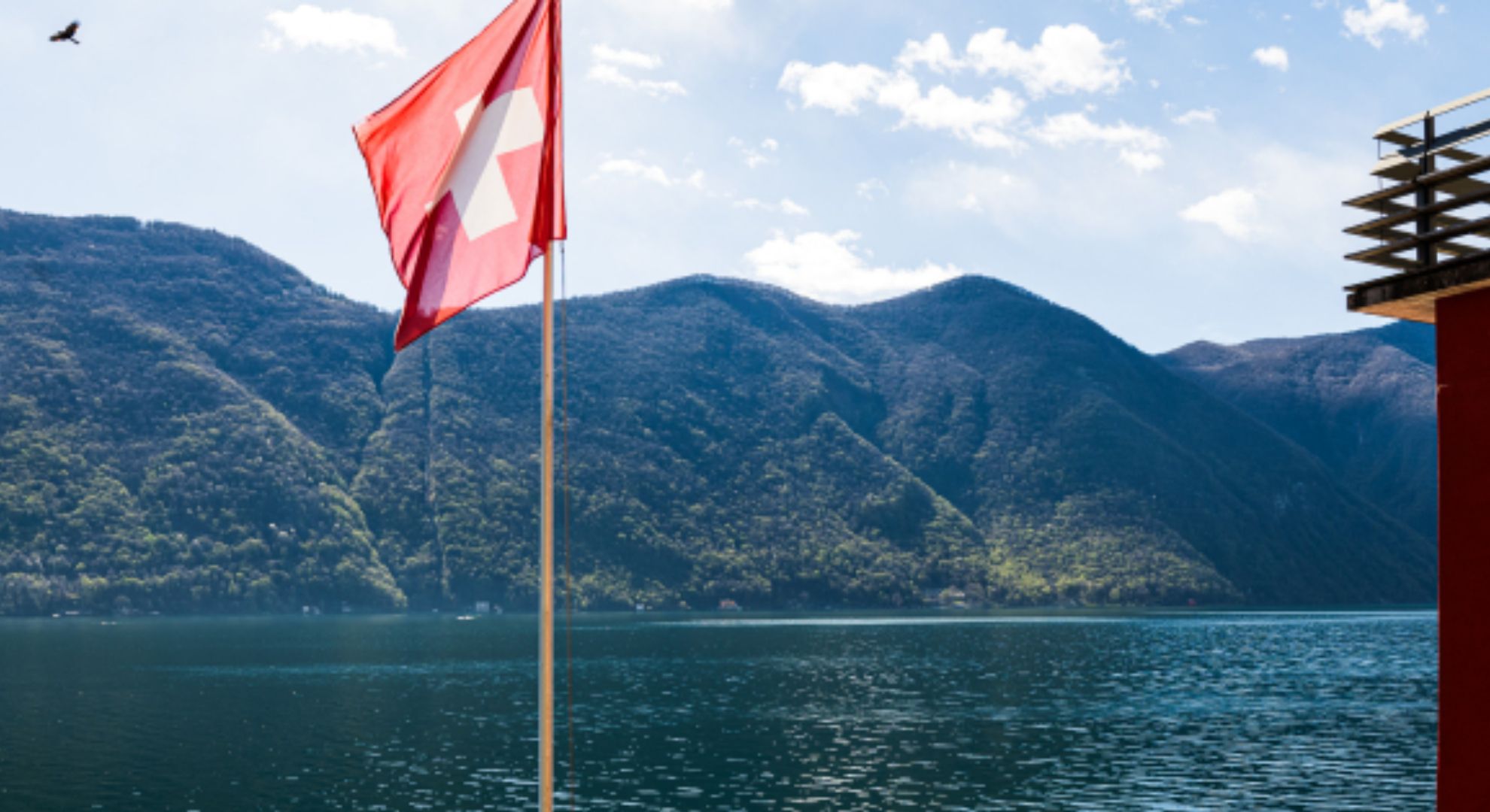 Bild des Luganersees bei schö-nem Wetter. Eine Schweizerfahne mit Mast ist sichtbar