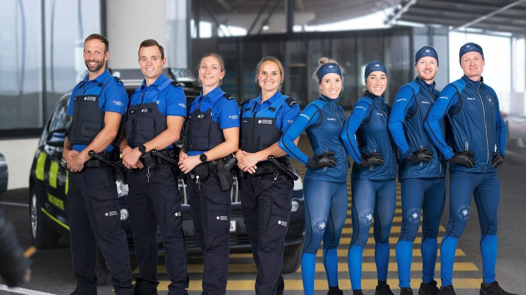 Gruppenfoto des Spitzensportteams des BAZG in blauen Langlauf- und Biathlonanzuegen vor hellem Hintergrund. Die Athletinnen und Athleten posieren mit Langlaufskis und Biathlongewehren in zwei Reihen.  