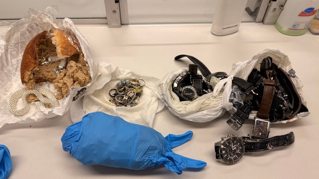 Verschiedene Schmuckstücke und Uhren, die in einem Brot und Plastiksäcken versteckt waren, sind auf einem Tisch ausgebreitet. Eine blaue Schutzhandschuhhand ist ebenfalls sichtbar.  