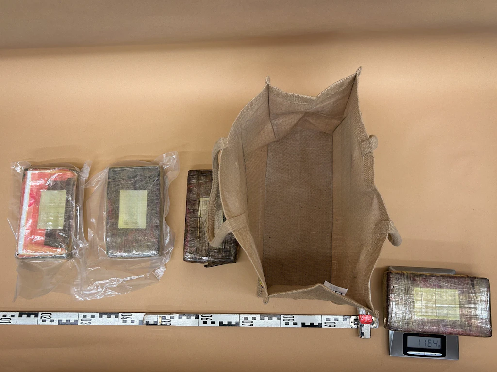Auf dem Bild sind mehrere in Plastik verpackte rechteckige Pakete zu sehen, die vermutlich Drogen enthalten. Daneben liegt eine leere Stofftasche sowie eine Waage, auf der eines der Pakete gewogen wird. Ein Massband dient zur Grösseneinschätzung.