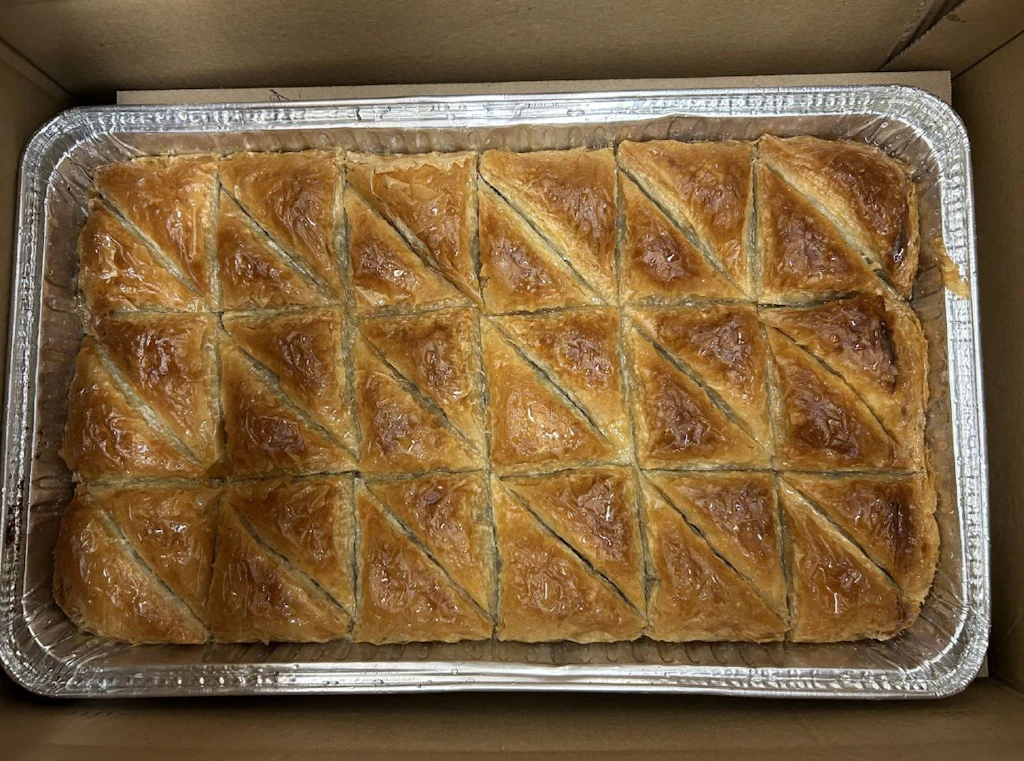 In einem Karton befindet sich ein grosses Blech mit frisch gebackenen, goldbraunen türkischen Backwaren, die in Dreiecke geschnitten sind. Die Oberfläche ist glänzend und zeigt die typische Schichtung von Blätterteig. 
