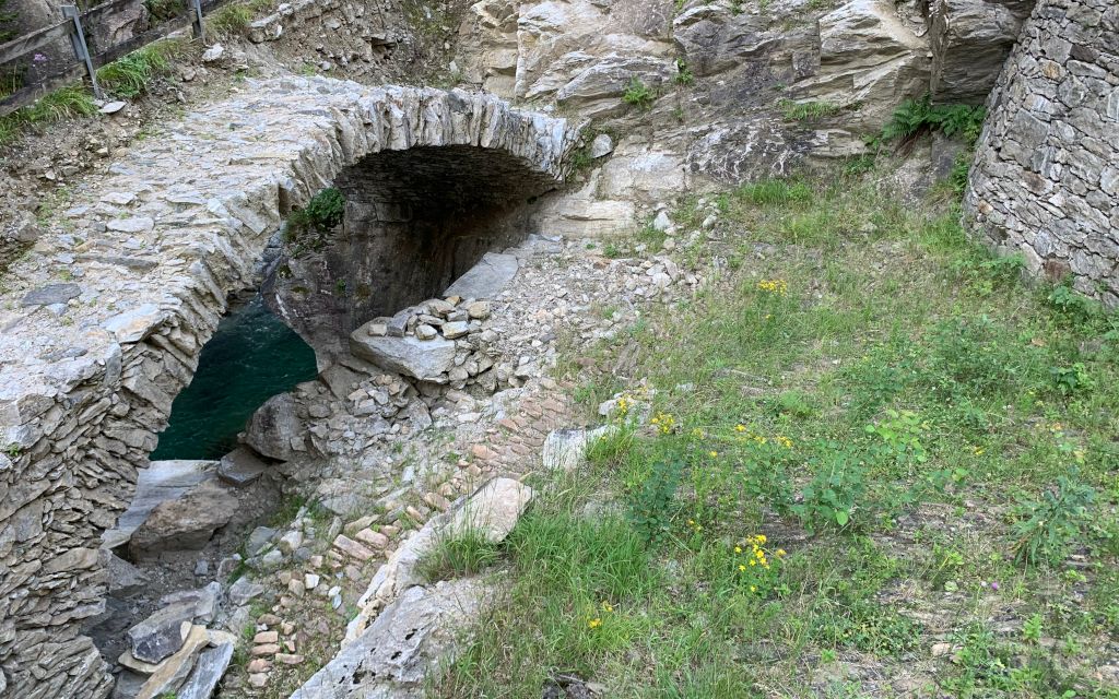 Eine alte Steinbruecke in einer ländlichen Umgebung, umgeben von Gras und Felsen. Die Bruecke ist teilweise verfallen und zeigt Spuren der Zeit.  