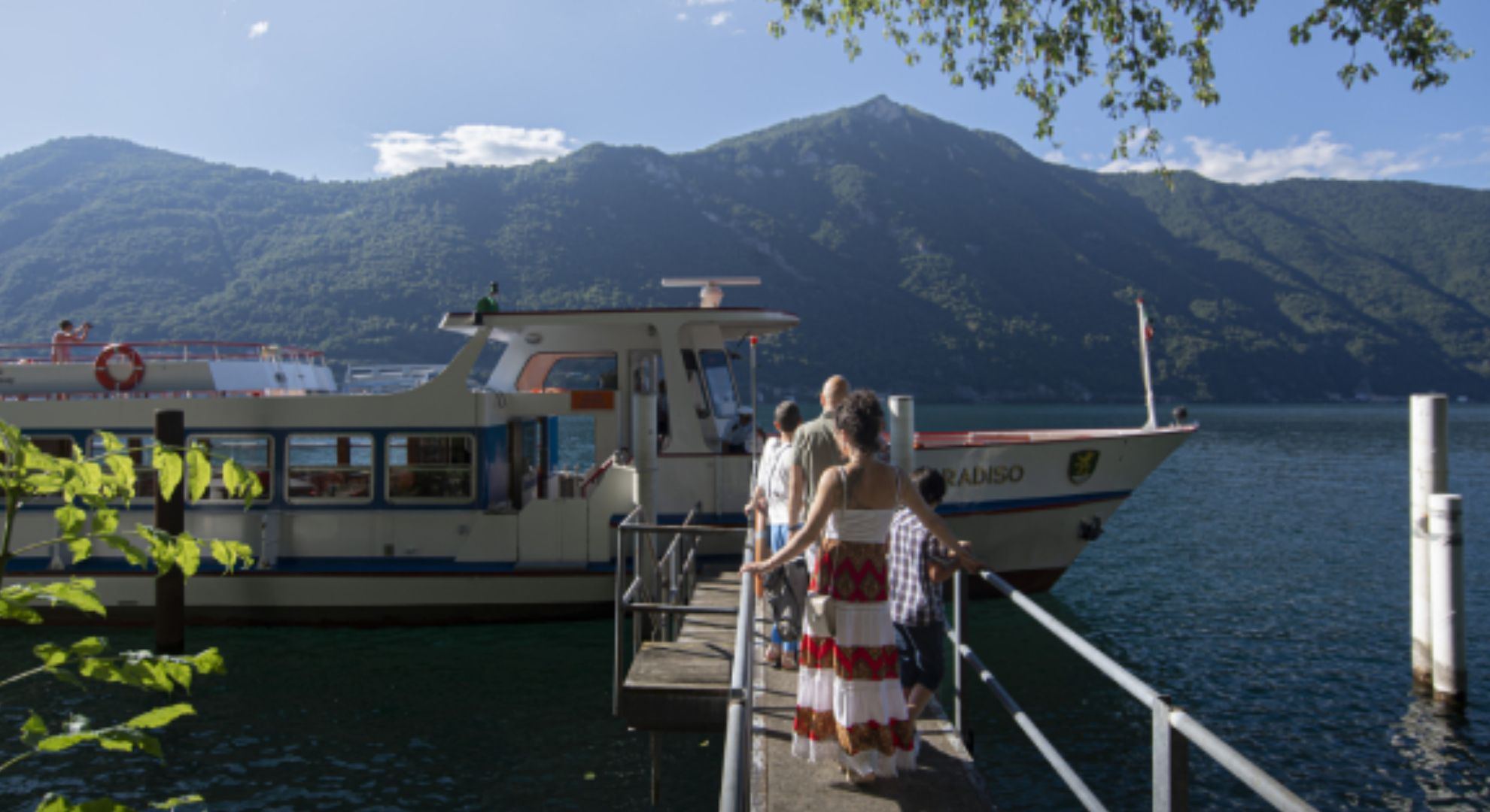 Personen stehen an der Schiffhal-testelle und warten, dass sie auf dem Schiff einsteigen können. 