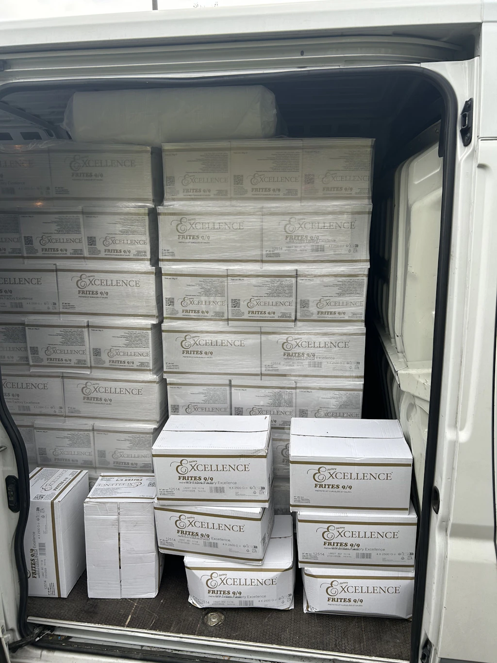 In einem weissen Lieferwagen sind zahlreiche Kartons der Marke Excellence Frites 9/9 gestapelt. Die Kartons sind ordentlich aufeinandergeschichtet und füllen den Laderaum fast vollständig aus. Es handelt sich offenbar um einen Warentransport.  