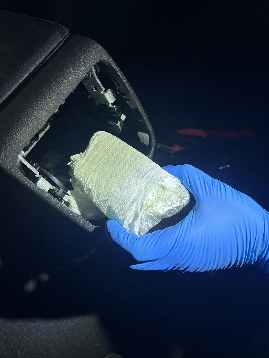 Ein Paar blaue Handschuhe hält ein in Plastik verpacktes Bündel aus einem Autoversteck im Dunkeln.