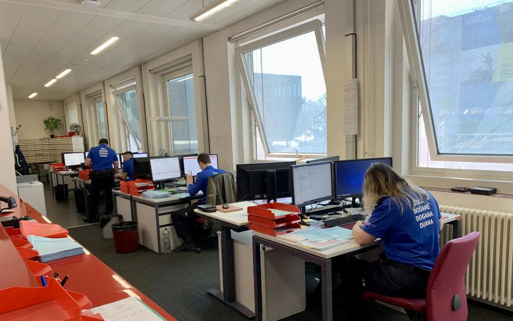 Mehrere Zollmitarbeitende sitzen in einem Buero an Computerarbeitsplaetzen und bearbeiten Dokumente. Sie tragen blaue T-Shirts mit der Aufschrift Zoll in verschiedenen Sprachen. Auf den Tischen liegen zahlreiche rote Ablageboxen und Papierstapel.  