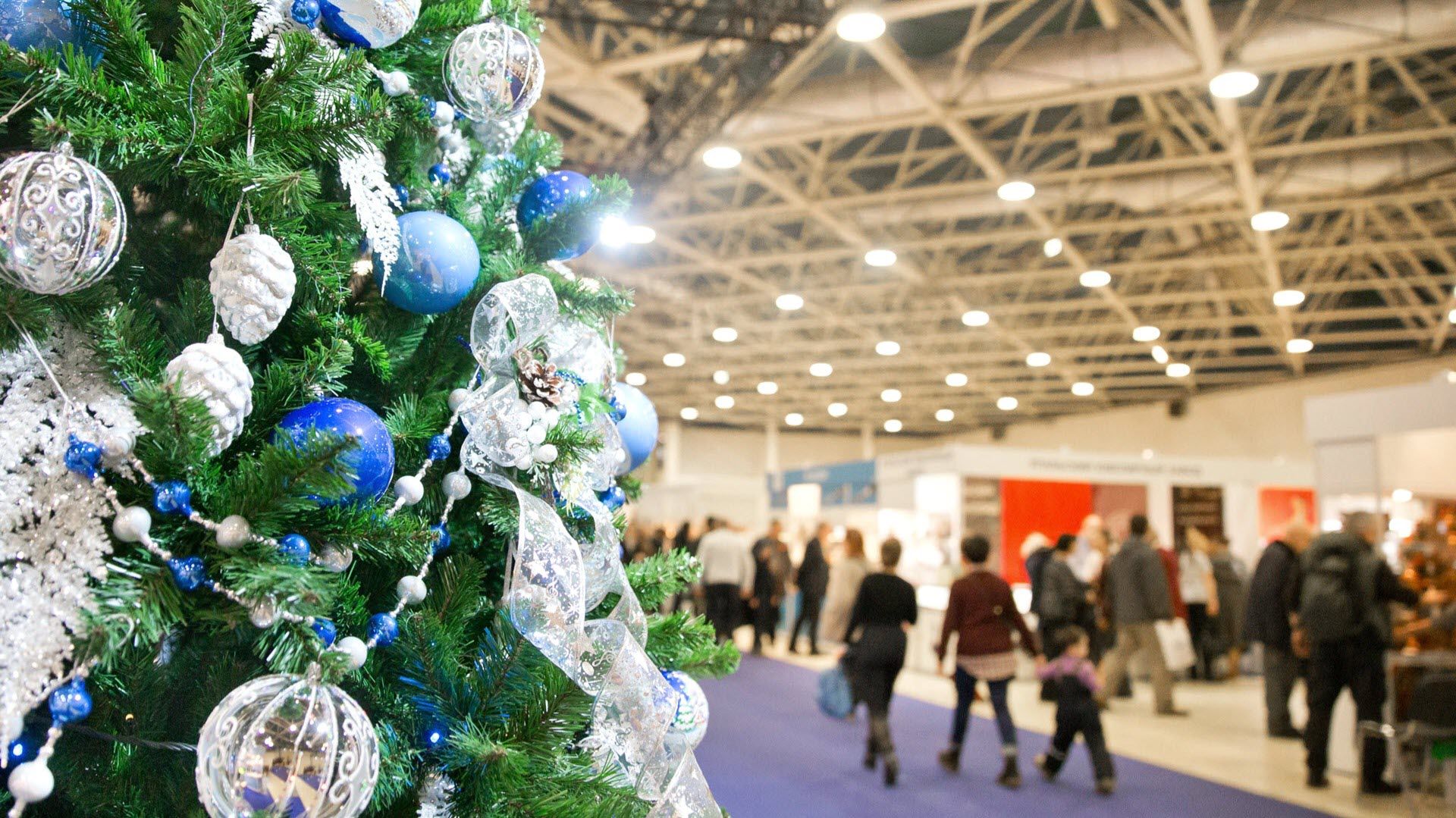 Ein geschmückter Weihnachtsbaum mit blauen und silbernen Kugeln steht in einem Kaufhaus. Im Hintergrund sind unscharf Menschen und Stände zu sehen