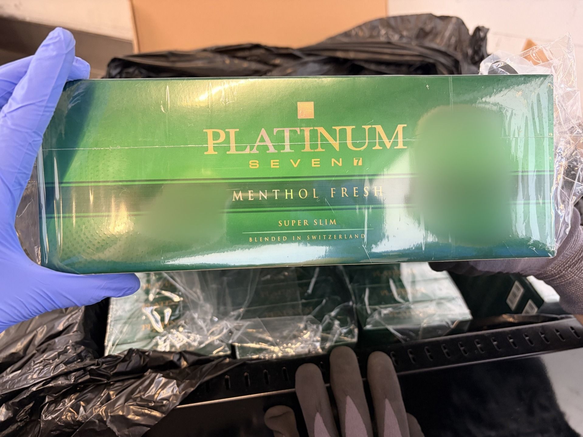 Eine Person mit Handschuhen hält eine Packung Platinum Seven Menthol Fresh Zigaretten, die in einem Computerrack versteckt wurden. Im Hintergrund sind weitere Zigarettenpackungen sichtbar. Die Szene deutet auf eine Kontrolle oder Beschlagnahmung hin.