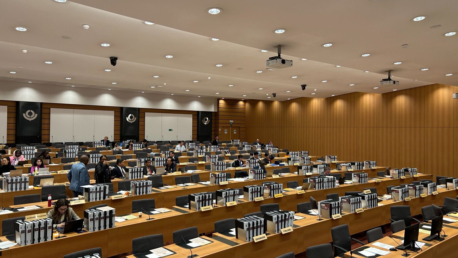 Ein großer Konferenzraum mit vielen Tischen, an denen Menschen sitzen. Auf den Tischen stehen zahlreiche Ordner.