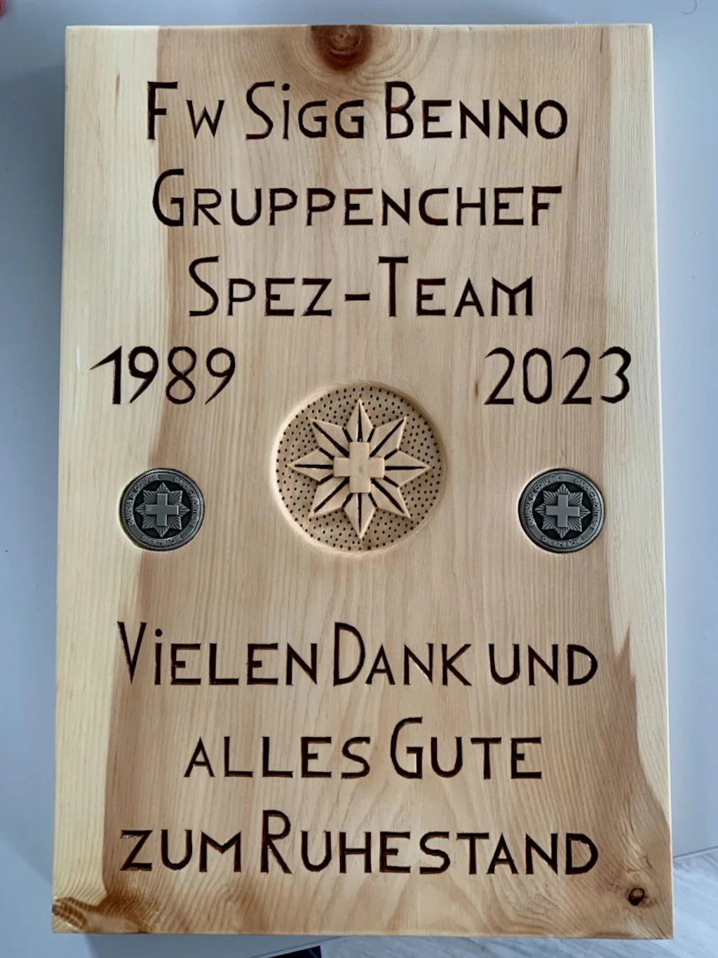 Holztafel mit eingraviertem Dankestext an den Gruppenchef des Spezial-Teams, den Jahreszahlen 1989 und 2023 sowie zwei Emblemen mit Schweizer Kreuz. In der Mitte ist ein sternförmiges Relief mit Kreuz zu sehen.