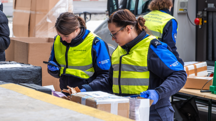 Zwei Frauen in Uniformen des BAZG arbeiten an einem Tisch mit Paketen. Eine Frau benutzt ein Smartphone, während die andere Pakete überprüft.