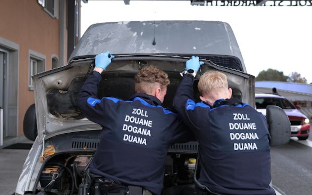 Zwei Zollbeamte inspizieren den Motorraum eines Fahrzeugs an einem Grenzübergang. Sie tragen blaue Uniformen mit der Aufschrift "Zoll" in mehreren Sprachen. 
