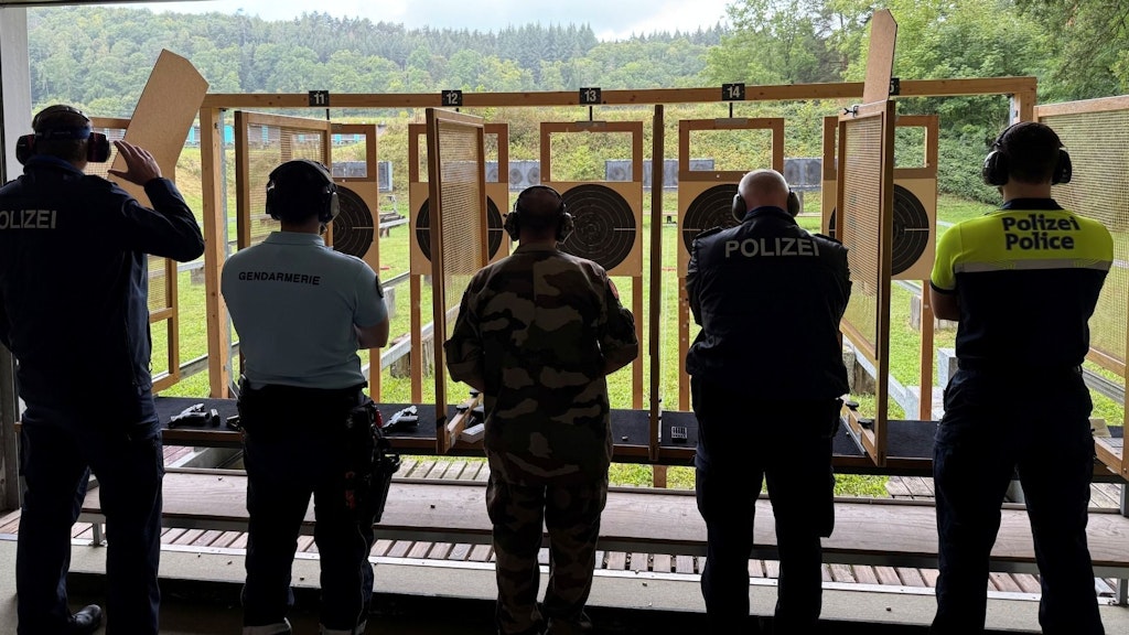 Fünf Personen in Uniform stehen vor Zielscheiben auf einem Schießstand im Freien. Sie tragen Gehörschutz und konzentrieren sich auf das Schießen. Die Umgebung ist grün und bewaldet.  