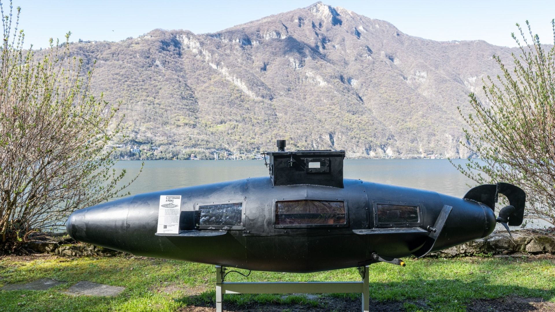Ein kleines, schwarzes U-Boot steht auf einem Gestell vor einem See mit Bergen im Hintergrund.  