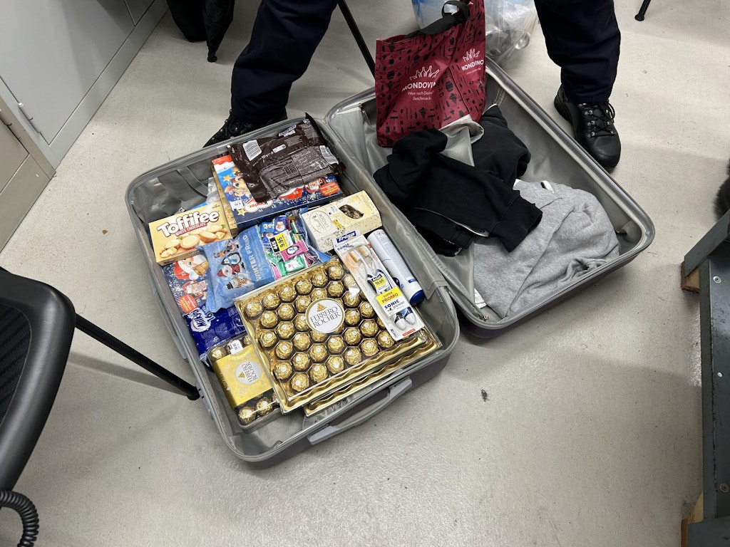 Offener Koffer mit Süssigkeiten und Kleidung in einem Raum mit linoleum Boden. Zwei Personen stehen dabei.