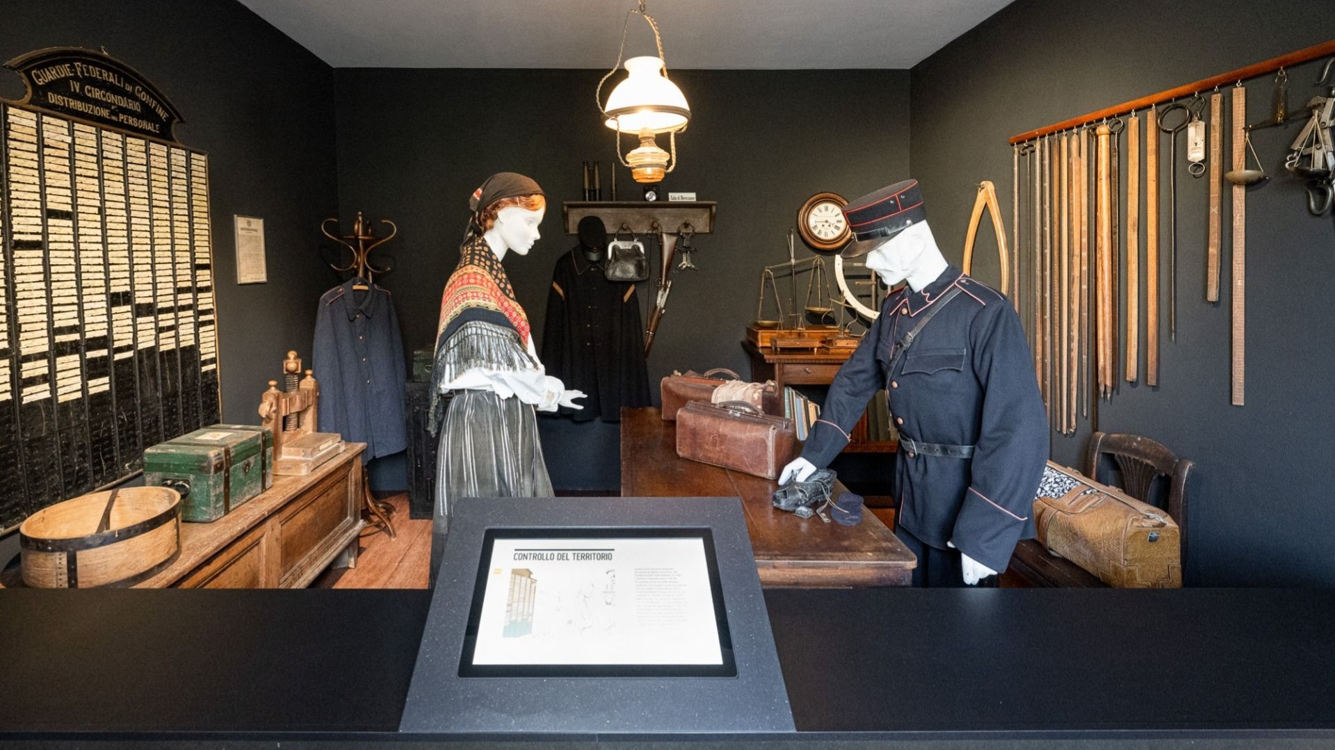 Eine historische Zollszene mit zwei Figuren in traditioneller Kleidung in einem alten Büro. Verschiedene antike Gegenstände und Werkzeuge sind im Raum verteilt.