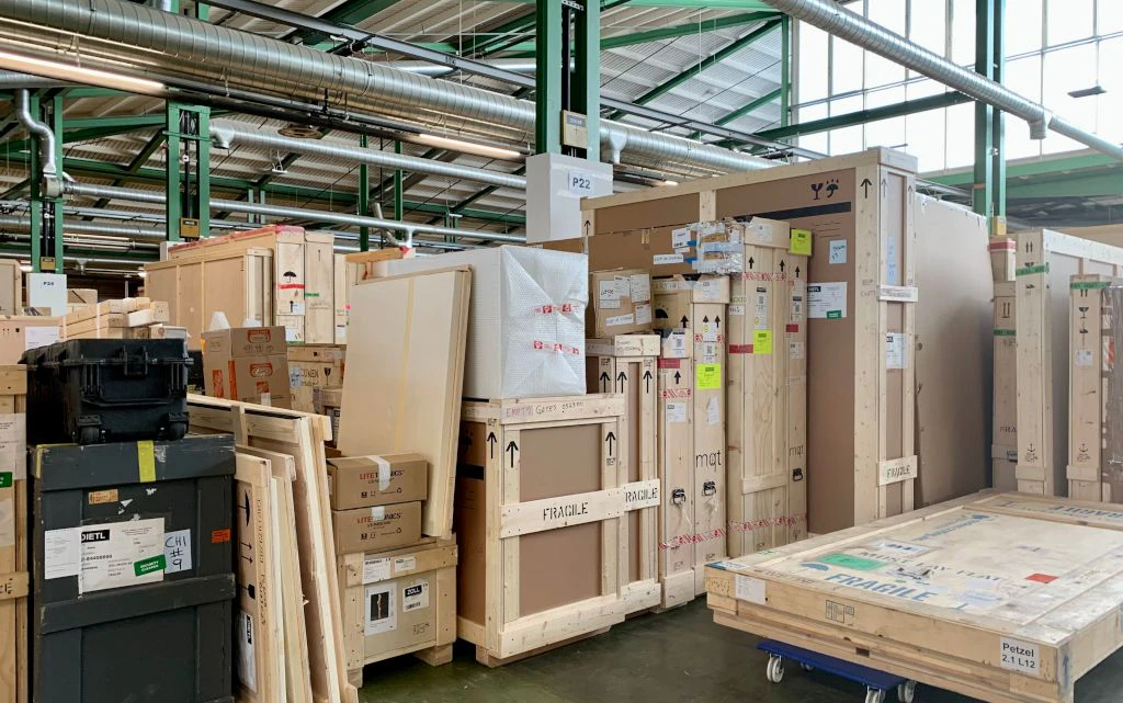 In einer Lagerhalle stehen zahlreiche Holz- und Kunststoffkisten dicht aneinandergereiht, viele davon mit Aufklebern und der Aufschrift "Fragile" versehen. Die Szene zeigt verpackte Sendungen, die offenbar für Transport oder Zollabfertigung bereitstehen.  