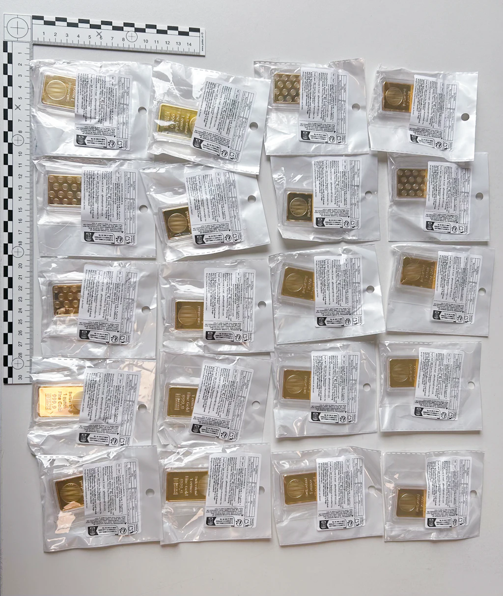Mehrere einzeln verpackte Goldbarren liegen auf einer weissen Fläche. Die Barren sind in transparenten Kunststoffhüllen mit Etiketten und Sicherheitsmerkmalen versiegelt. Ein Massstab zur Grösseneinschätzung ist links oben sichtbar.