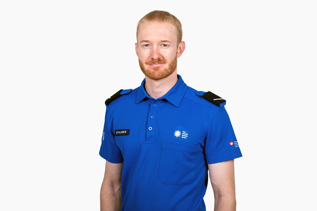 Ein Mitarbeiter des BAZG in blauer Uniform steht vor einem weissen Hintergrund. Auf seiner Uniform sind verschiedene Abzeichen sichtbar. Der Mitarbeiter hat kurzes, blondes Haar und einen Bart.