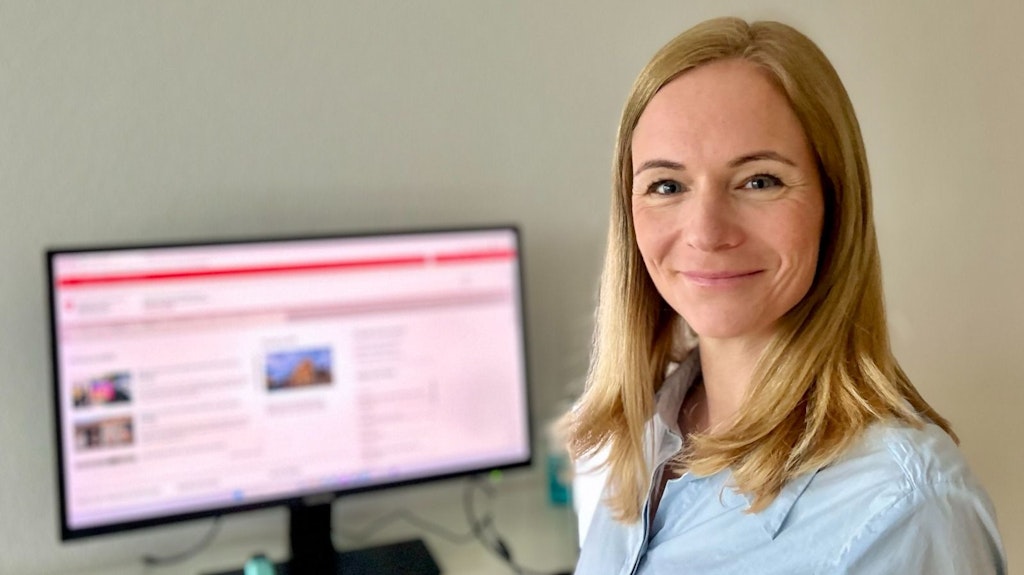 Eine Frau steht lächelnd vor einem Computerbildschirm. Der Bildschirm zeigt eine unscharfe Webseite.  