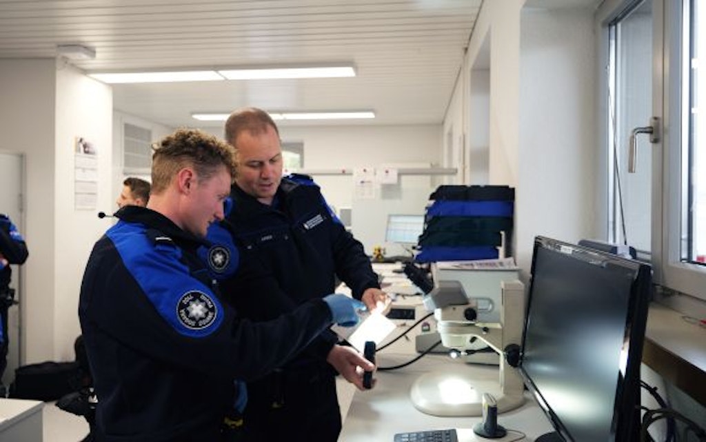 Zwei Zollbeamte in Uniform inspizieren ein Dokument in einem Büro. Sie stehen vor einem Computer und einem Mikroskop.