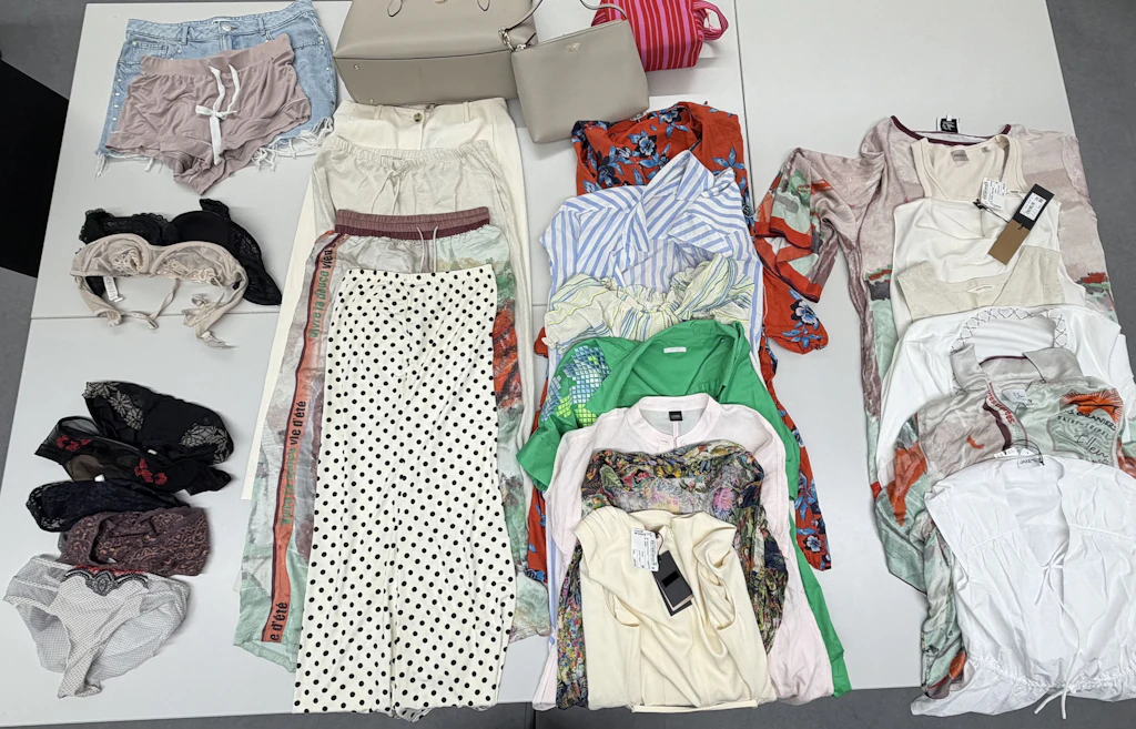 Verschiedene Kleidungsstücke, Unterwäsche und Accessoires wie Taschen liegen ordentlich auf einem Tisch ausgebreitet. Die Auswahl umfasst Hosen, Shorts, Oberteile, Kleider und Taschen in unterschiedlichen Farben und Mustern. Die Artikel wirken neu und teilweise mit Etiketten versehen. 