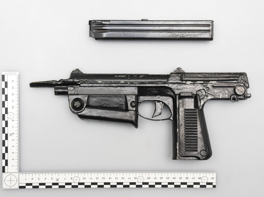 Eine schwarze Pistole und Magazin auf weissem Hintergrund, mit Massstab daneben. Die Pistole zeigt Abnutzung und ist auf der linken Seite platziert.